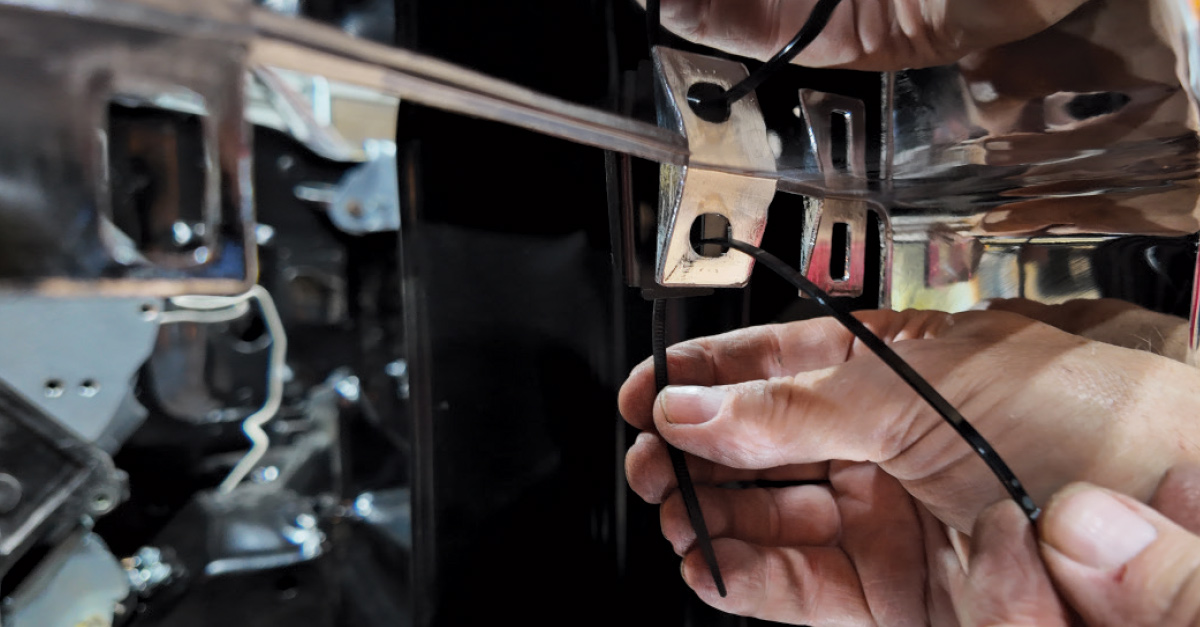 Close-up of hands using black zip ties to secure components behind a chrome car part.