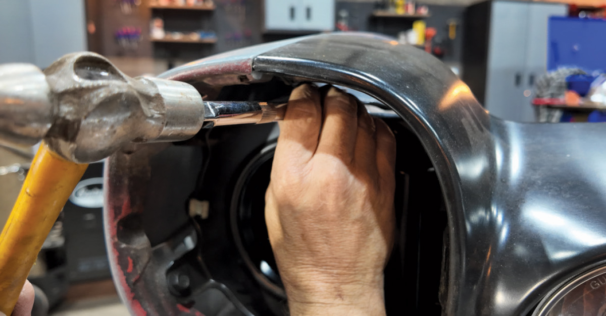 Hands using a tool and hammer to adjust the alignment of a black car body panel.