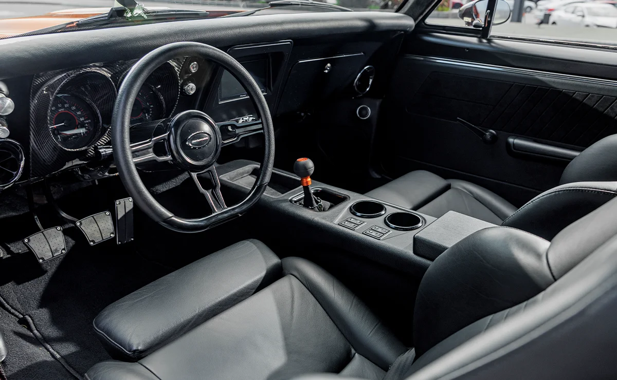 A close-up view of a modified classic car cockpit with carbon fiber dashboard accents, aftermarket gauges, a digital touchscreen display, and integrated cup holders in a sleek black leather console