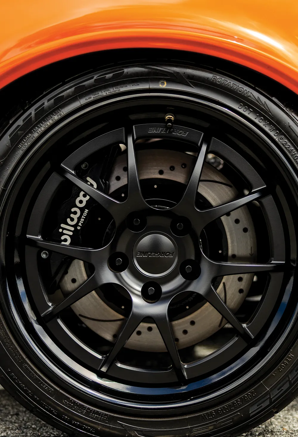 Close-up of a Forgeline matte black multi-spoke racing wheel on a customized orange muscle car, featuring a high-performance Wilwood 6-piston brake caliper and slotted rotors