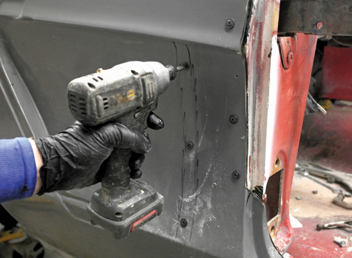 A gloved hand uses a power drill to drive a screw into a grey fiberglass body panel, with Sharpie marker markings visible on the surface