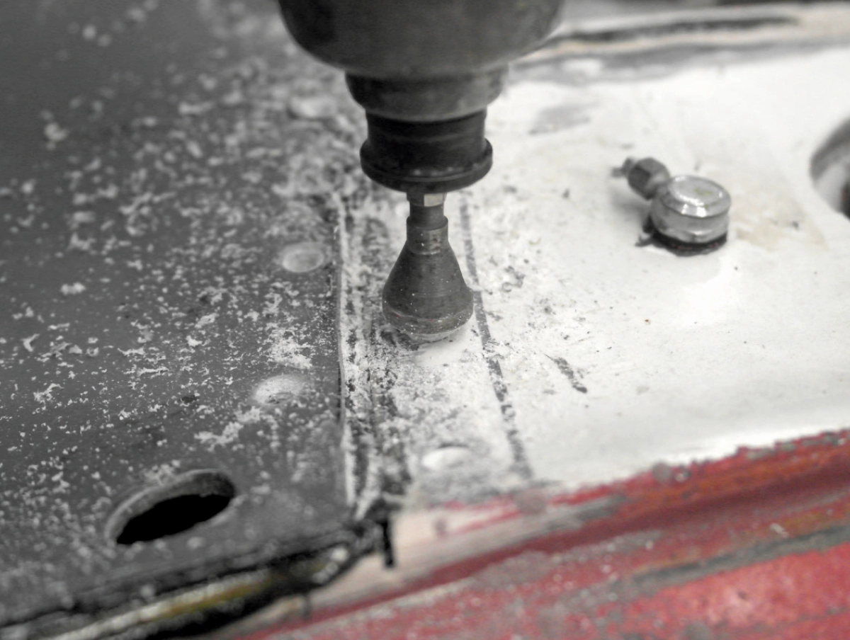 A close-up of a drill bit creating a countersink hole on the edge of the grey fiberglass panel, with debris scattered on the surface