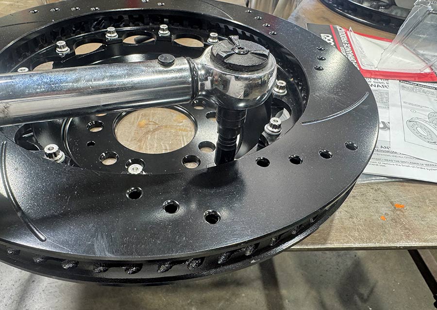 Mechanic using a torque wrench to secure bolts on a multi-piece, black, cross-drilled, and slotted high-performance brake rotor. Numerous 12-point bolts connect the rotor ring to its central hub