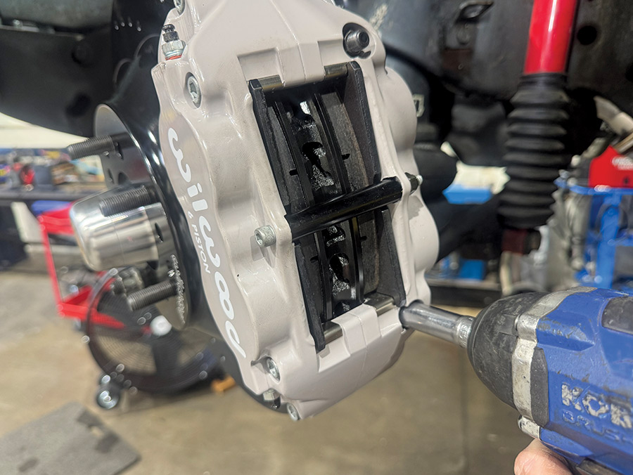 A blue cordless impact wrench is used to tighten the mounting bolts of a grey Wilwood 6-piston brake caliper over a black high-performance rotor