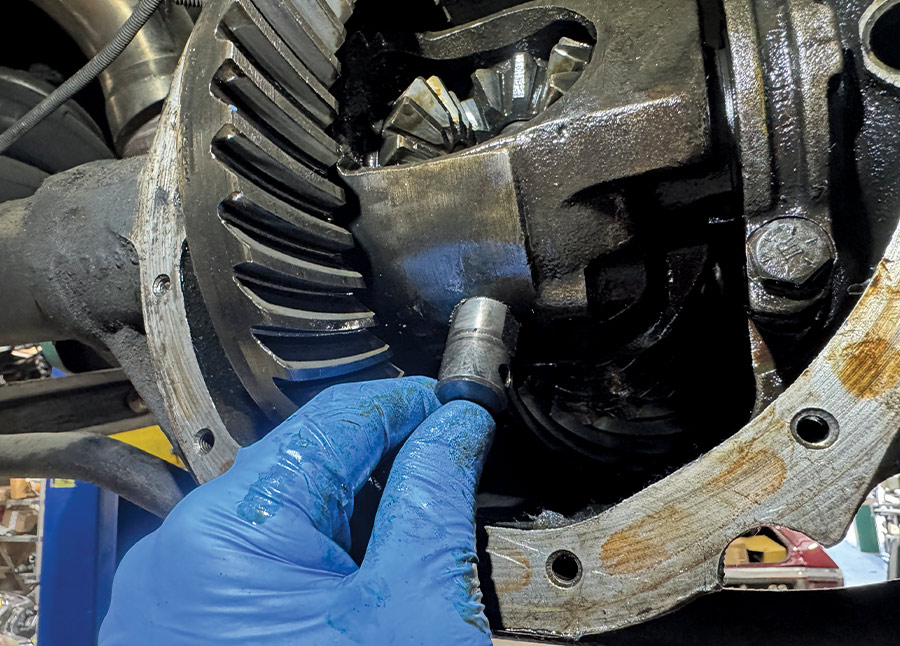 Close-up of a blue-gloved hand holding a metal cross-pin removed from a car's rear differential, with internal spider gears and the ring gear exposed