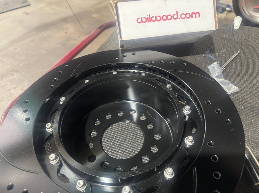 A black Wilwood multi-piece brake rotor with cross-drilled and slotted surfaces sits on a workbench next to its original packaging