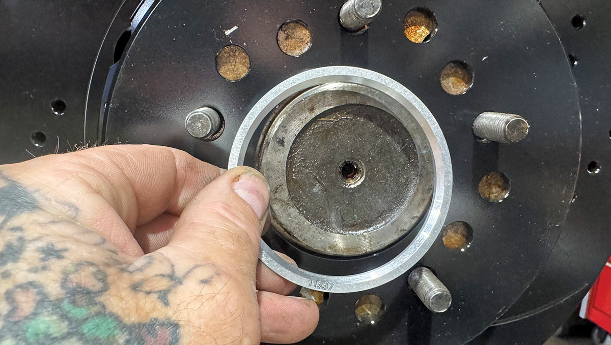 A tattooed hand installs a thin silver hub-centric ring, etched with the number 11337, onto the center of a black brake rotor hub