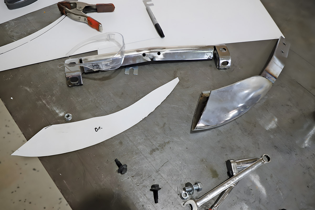 Chrome bumper sections, a white paper template for a bumper strake, and various hand tools laid out on a metal workbench.