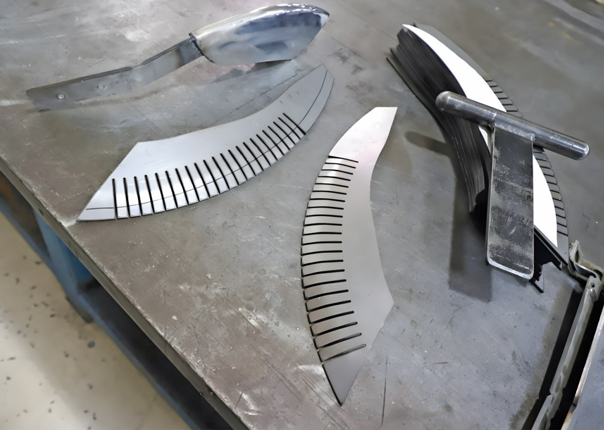 Steel bumper strake blanks with vertical relief cuts for forming curves, shown next to a metal fabrication tool on a workshop table.