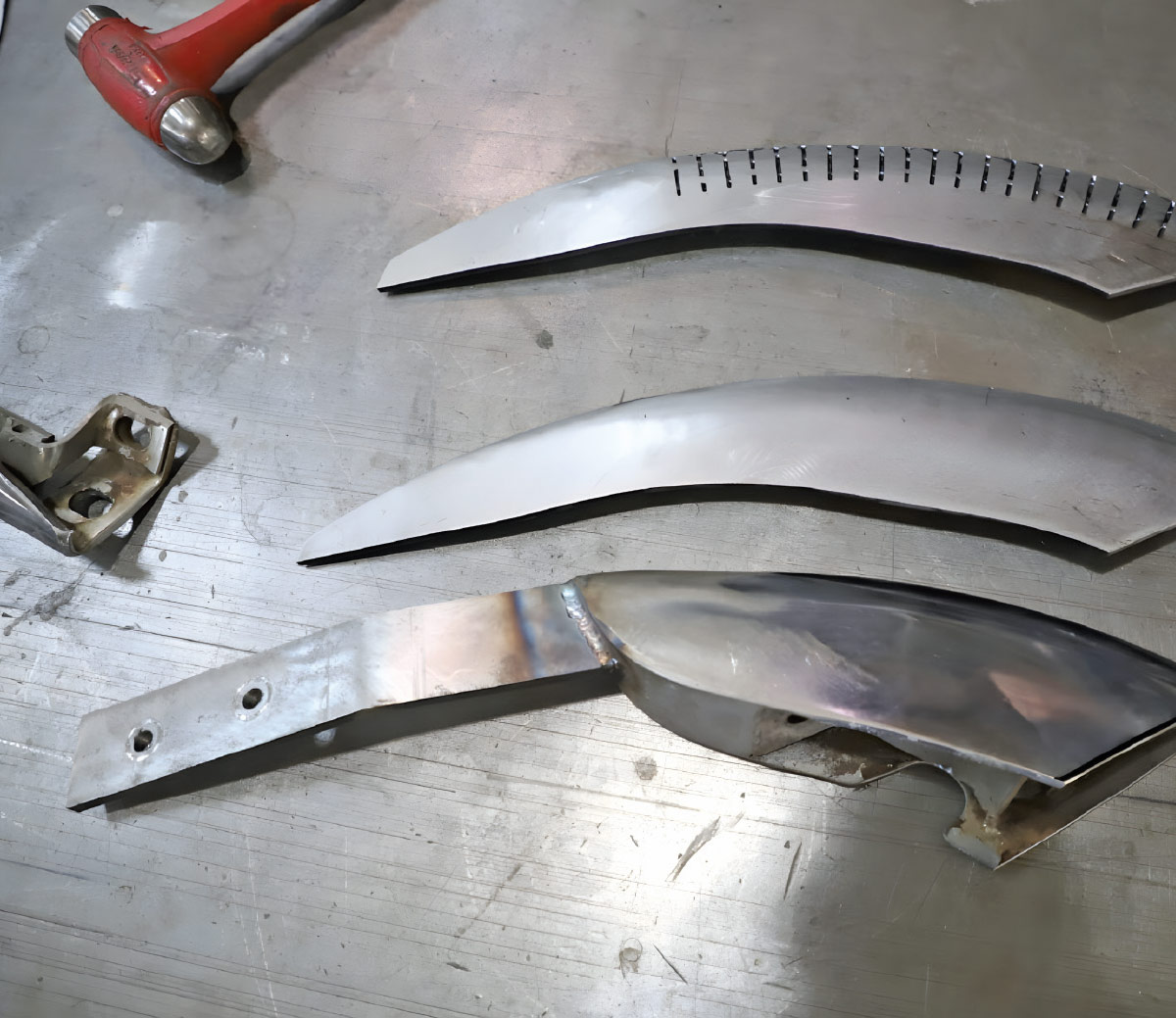 Three metal bumper strakes on a workbench next to a red hammer. The pieces show stages of fabrication: a sliced metal blank, a shaped replacement, and an original factory strake.
