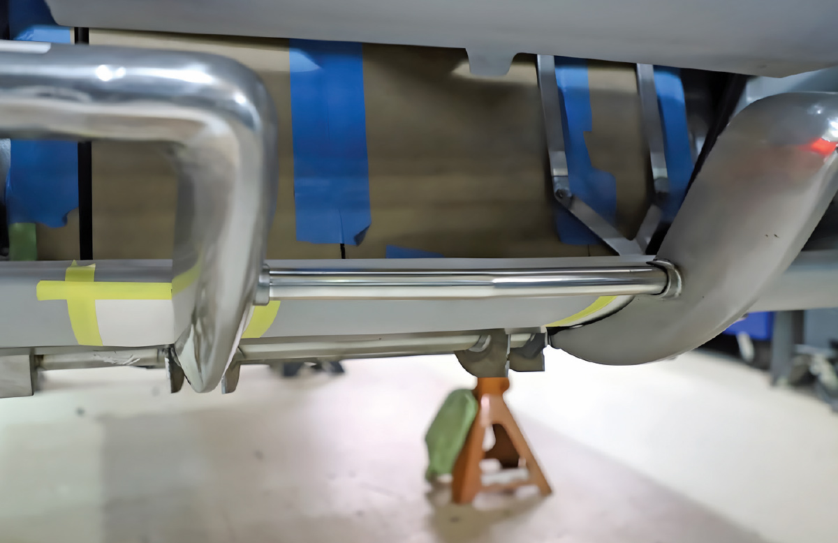 Close-up from below of a front bumper under fabrication. A metal spreader bar is fitted between the left and right curved bumper halves, which are held with masking tape.