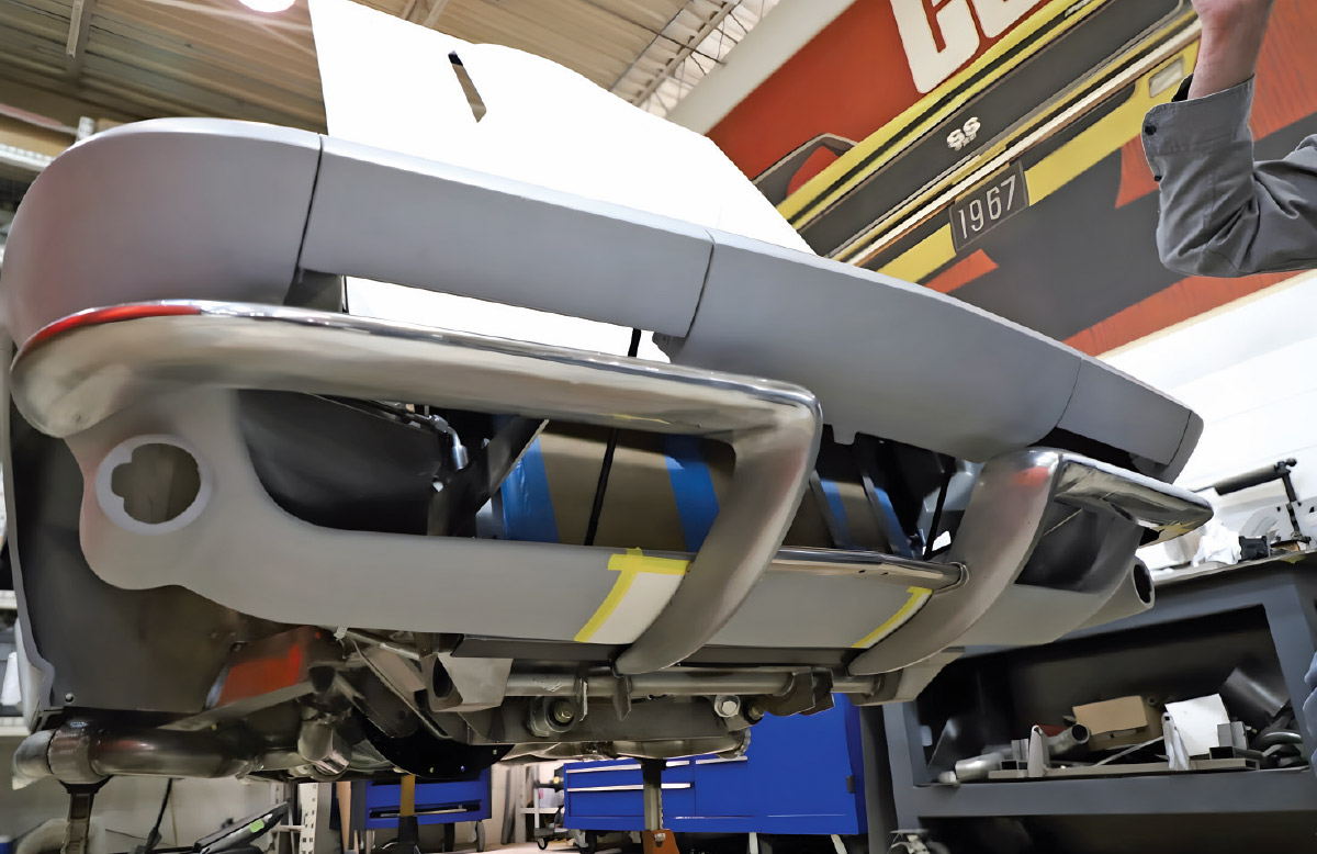 Low-angle view looking up at the front of a C2 Corvette in primer grey. The reshaped chrome front bumper, complete with new vertical strakes and spreader bar, is fitted, following the body contours.