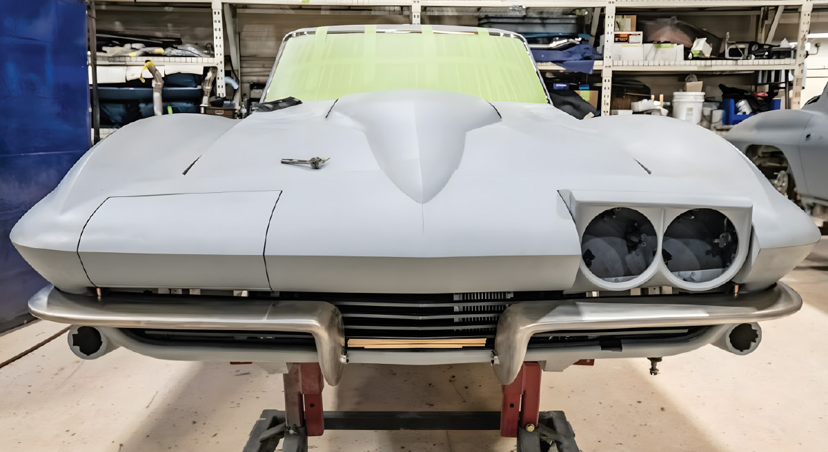 Straight-on front view of the primed C2 Corvette on a frame machine. The green masked windshield area and the new fabricated bumper are visible. One headlight door is open.