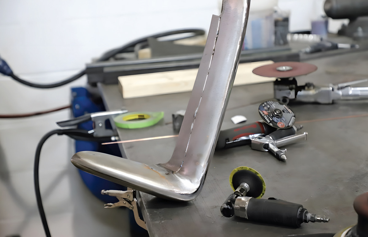 A metal bumper section stands on a workbench with a thin metal strip welded to its edge. Pneumatic grinders and shop tools surround the piece.