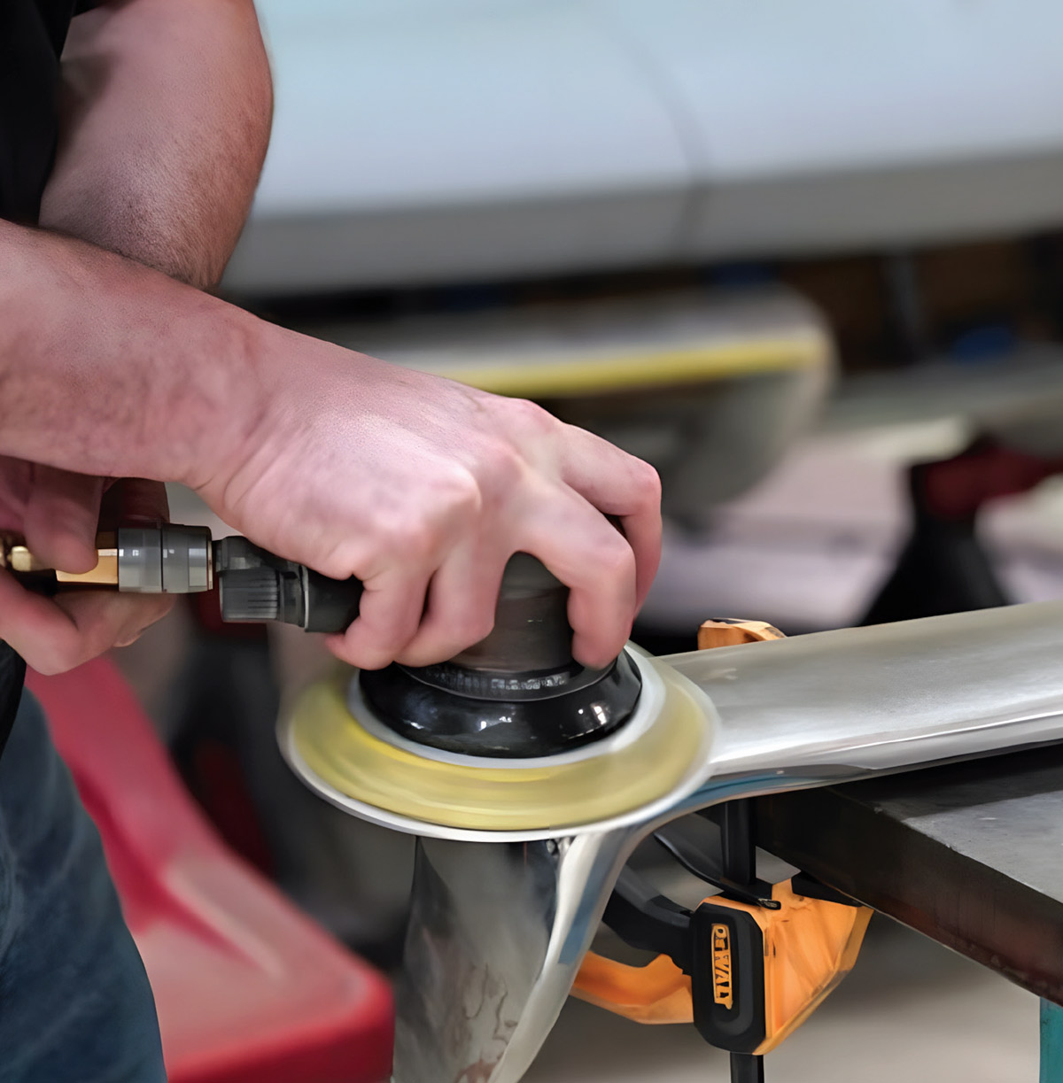 Close-up of hands operating a pneumatic orbital sander on a metal bumper part. The component is secured to a metal workbench with a yellow plastic clamp.