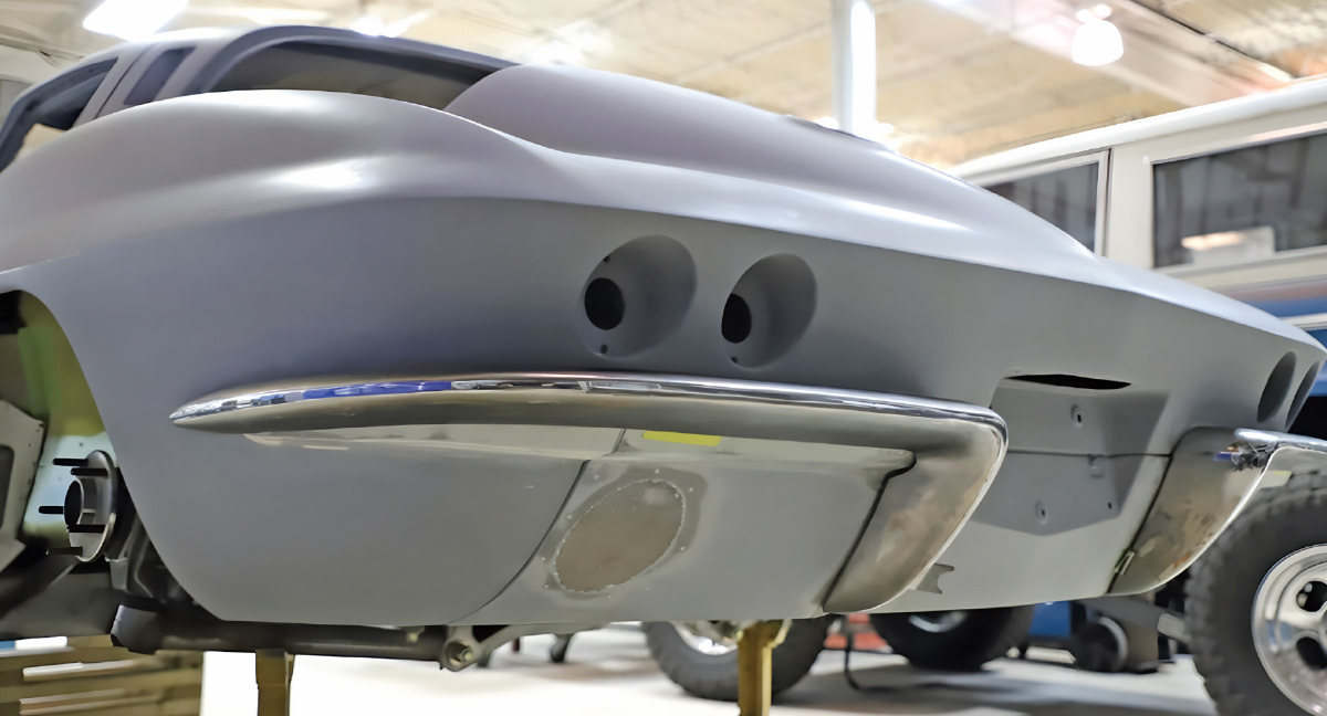 Straight-on rear view of a grey primed C2 Corvette with a fabricated metal bumper installed. The bumper follows the lower body contour beneath two empty circular taillight housings.