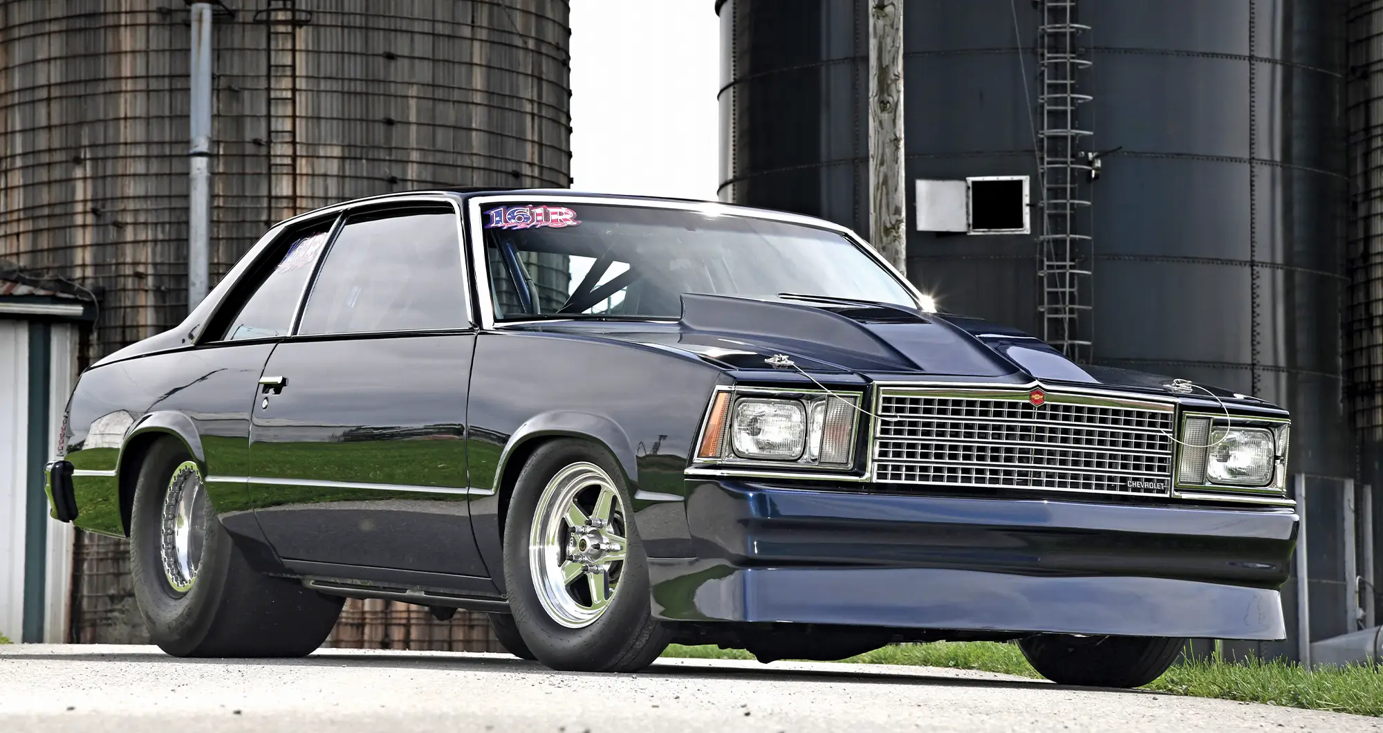 Front three-quarter view of a dark blue Chevy Malibu with a cowl hood, hood pins, and a deep front spoiler, parked in front of two large farm silos.