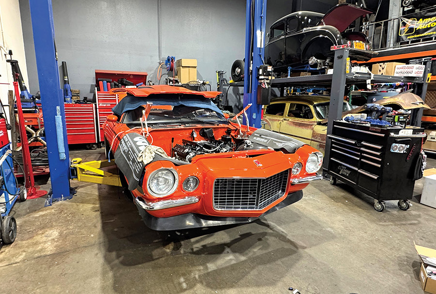 An orange classic muscle car sits on a lift in a busy automotive restoration shop.