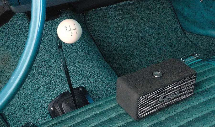 A classic car interior featuring teal carpeting, a chrome manual shifter with a white four-speed knob, and a black Marshall portable speaker resting on the striped teal seat.